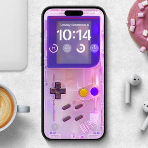 Puede incluir: Un smartphone con una funda rosa transparente con un diseño de videojuego retro. La pantalla muestra la hora, la fecha e iconos. En la imagen también hay un donut rosa glaseado con malvaviscos, una taza de café y auriculares blancos.
