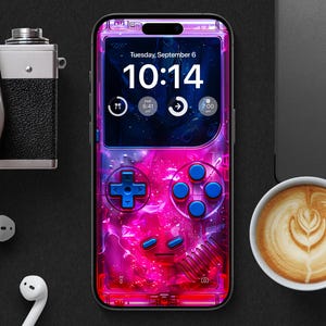 Puede incluir: Un smartphone con un diseño retro de videojuego rosa y azul, que muestra la hora 10:14 en la pantalla. La imagen incluye una cámara, auriculares y una taza de café con arte latte, todo sobre una superficie negra.