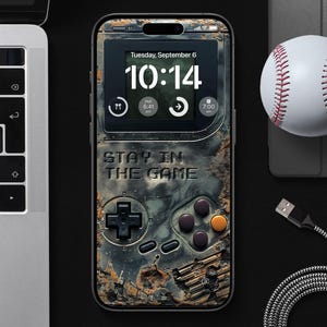 以下が含まれることがあります： 黒と緑の携帯電話ケースで、すり切れて古びたような外観です。ケースには「10:14」と表示されたデジタル時計と「STAY IN THE GAME」という文字があります。ケースは、ボタンとジョイスティックが付いた、レトロなビデオゲーム機のようなデザインになっています。