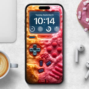以下が含まれることがあります： ピンクとオレンジのふわふわのケースをつけたスマートフォン。電話の画面には、10:14 の時刻と、9 月 6 日火曜日の日付が表示されています。ケースは、ボタンと方向キーが付いたレトロなビデオゲームコントローラーのデザインになっています。