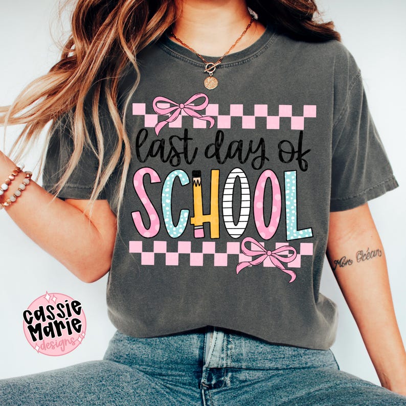May include: Dark gray t-shirt with the text "last day of SCHOOL" in colorful letters. The design includes pink bows and a pink and white checkered pattern. The shirt is a casual, short-sleeved style.