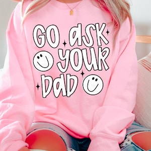 Puede incluir: Sudadera rosa con el texto "GO ASK YOUR DAD" en un diseño en blanco y negro. El diseño incluye caras sonrientes y detalles de estrellas. La sudadera es de color rosa liso.