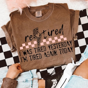 May include: A brown Comfort Colors t-shirt with the word "retired" in black script, a lightning bolt graphic, and the text "I WAS TIRED YESTERDAY I'M TIRED AGAIN TODAY" in black. The shirt has rolled-up sleeves.