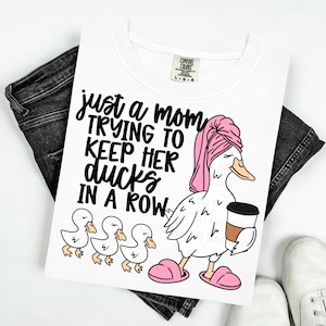 May include: White t-shirt with the text "just a mom trying to keep her ducks in a row." The graphic features a cartoon duck wearing a pink towel and slippers, holding a coffee cup, with three ducklings.