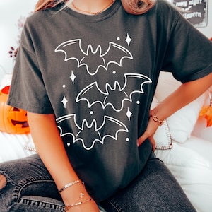 May include: Dark gray t-shirt featuring a white graphic of three bats and sparkling stars. The shirt is a comfortable, casual style, perfect for Halloween or spooky season. The design is simple and eye-catching.