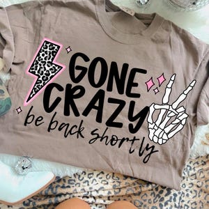 May include: A taupe-colored t-shirt with the words "GONE CRAZY" and "be back shortly" in black script. A pink-outlined lightning bolt with a leopard print and a skeleton hand making a peace sign are also featured.