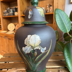 May include: A dark gray ceramic jar with a lid, featuring a hand-painted white iris flower with blue accents. The lid is green with a butterfly and leaf design. The jar has a green base and lid trim.