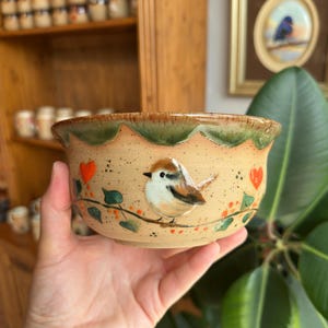Puede incluir: Un cuenco de cerámica con un diseño de pájaro pintado a mano. El cuenco tiene una base beige con un borde verde y marrón, y un pájaro pintado con corazones rojos y hojas verdes. El cuenco se sostiene en una mano.