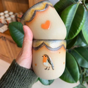 Puede incluir: Dos tazas de cerámica apiladas, sostenidas por una persona. Las tazas son beige con un borde festoneado en marrón y dorado. Una tiene un corazón naranja, la otra una ilustración de petirrojo. Pequeños corazones naranjas dispersos.