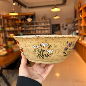 Op de afbeelding: Een handbeschilderde keramische kom met een gespikkelde beige buitenkant. De kom heeft een bloemenontwerp van witte madeliefjes, rode bessen en blauwe bloemen. De kom wordt voor een vage achtergrond gehouden.