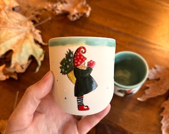 May include: A white ceramic mug with a hand-painted illustration of a gnome holding a Christmas tree. The gnome wears a red hat and black coat. The mug has a light blue rim and is next to a smaller green mug.