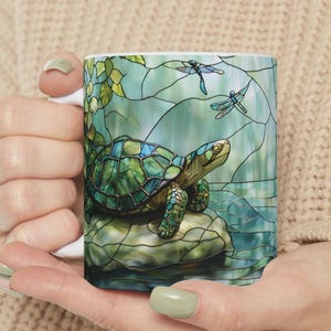 May include: White ceramic mug featuring a stained-glass style design of a turtle on a rock, with dragonflies and aquatic plants. The turtle has a green and blue shell. The background is a mix of blues and greens.