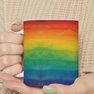 Può includere: Una tazza di ceramica bianca con un vivace design ad acquerello con bande orizzontali di rosso, arancione, giallo, verde, blu e viola. La tazza è tenuta tra le mani di una persona.