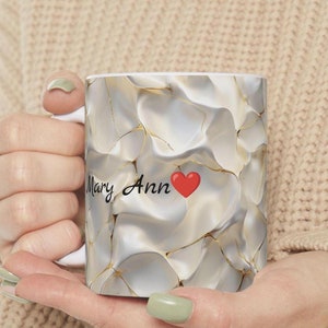 Puede incluir: Taza de cerámica blanca con un diseño de mármol dorado y blanco. La taza tiene el nombre "Mary Ann" escrito con un corazón rojo.