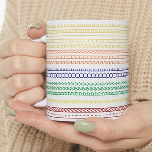 May include: White ceramic mug featuring a colorful pattern of horizontal lines in various designs and colors, including yellow, orange, blue, and green. The mug is held by a person wearing a beige sweater.