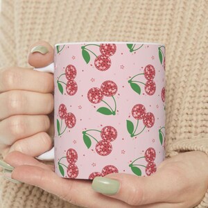 May include: A white ceramic mug with a pink background and a repeating pattern of red cherries with a disco ball design. The cherries have green stems and leaves.