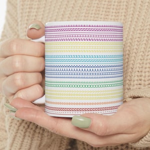 Pode incluir: Caneca de cerâmica branca com um padrão de arco-íris de vários desenhos costurados. O design da caneca apresenta faixas horizontais de vermelho, laranja, amarelo, verde, azul e roxo, cada uma com um padrão único semelhante a uma costura. A caneca tem uma alça branca.