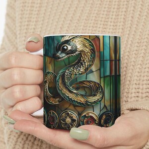 Könnte beinhalten: Eine weiße Keramiktasse mit einem farbenfrohen Design, das eine grüne und goldene Schlange mit einem Glasmalereieffekt zeigt. Die Schlange ist zusammengerollt und hat ein detailliertes Muster.