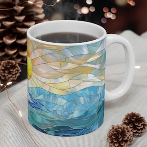May include: White ceramic mug with a stained glass design depicting a yellow sun over blue ocean waves. The mug has a white handle and is filled with a dark liquid. Decorative pinecones and string lights are in the background.