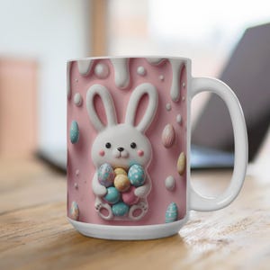 May include: A white ceramic mug with a pink background and a 3D bunny holding colorful Easter eggs. The bunny is white with pink ears and nose. The mug has a white handle.