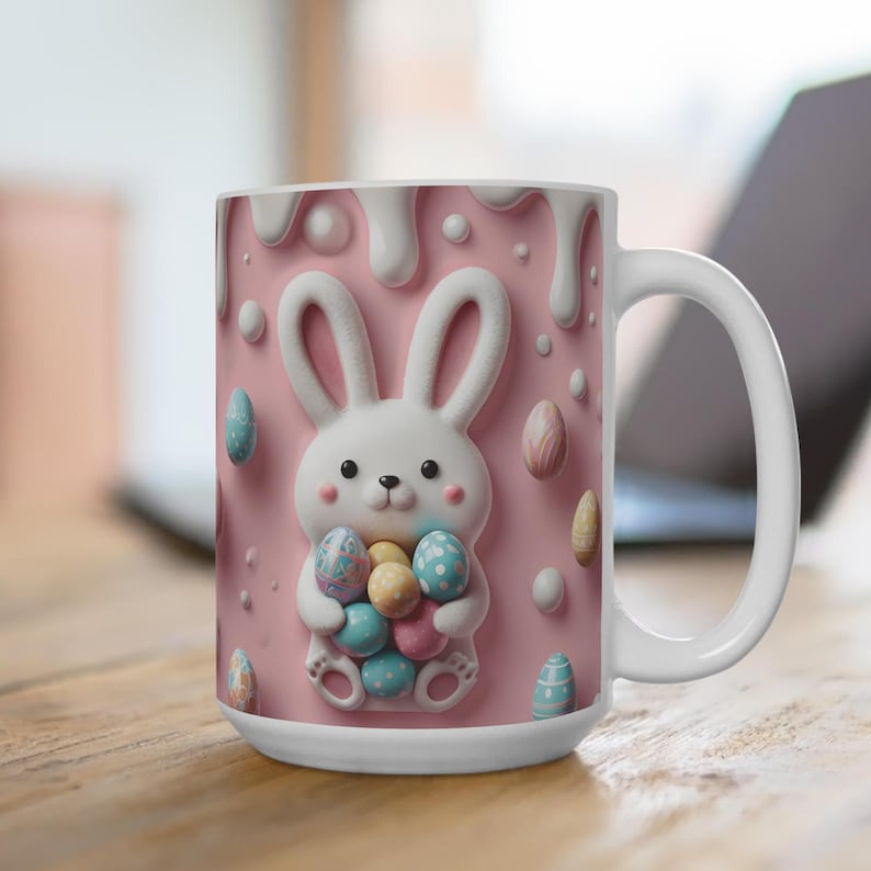 May include: A white ceramic mug with a pink background and a 3D bunny holding colorful Easter eggs. The bunny is white with pink ears and nose. The mug has a white handle.