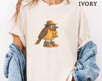 May include: Ivory-colored t-shirt featuring a cartoon bird wearing a hat, binoculars, and boots. The bird has brown and yellow feathers. The text "color: IVORY" is displayed on the upper right side of the shirt.