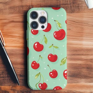 Puede incluir: Una funda para teléfono de color verde menta con un patrón de cerezas rojas. La funda tiene un recorte para el objetivo de la cámara y un borde blanco.
