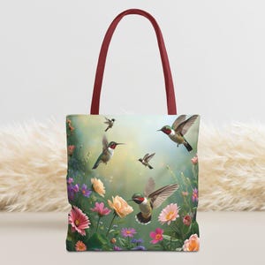May include: A tote bag featuring a vibrant design of hummingbirds in flight amidst colorful flowers. The bag has a burgundy handle and a soft, floral background. The design includes pink, yellow, and purple flowers.