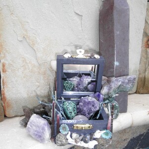 May include: A small wooden box with a glass lid filled with various crystals and stones, including amethyst, green aventurine, and a clear quartz sphere. The box is decorated with seashells and other natural elements. A tall, dark grey stone obelisk stands behind the box.
