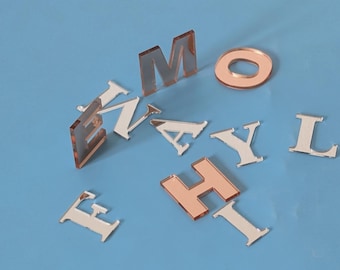 May include: A collection of silver and rose gold acrylic letters are arranged on a blue background. The letters include E, N, M, A, Y, L, H, and I, plus a rose gold circle. The letters are reflective and appear three-dimensional.