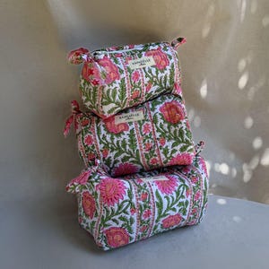 Peut inclure: Une pile de trois trousses matelassées rectangulaires de tailles différentes, présentant une base en tissu blanc avec un motif répétitif de fleurs roses et de feuilles vertes. Chaque trousse porte une étiquette avec le texte RAPHÄELLE.