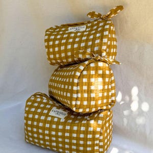Peut inclure: Une pile de trois trousses rectangulaires matelassées à motif vichy jaune moutarde et blanc. Chaque trousse présente un lien de fermeture en tissu sur le côté et une étiquette rectangulaire blanche avec le texte RAPHAELLE collection en noir.