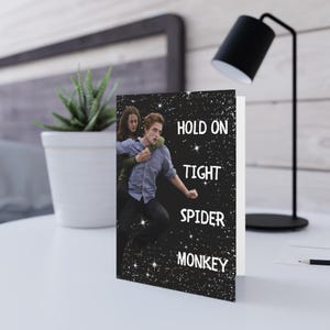 May include: A folded greeting card featuring a black background with white star-like sparkles. The front displays a photographic image of two people, one carrying the other on their back, with the white text "HOLD ON TIGHT SPIDER MONKEY".