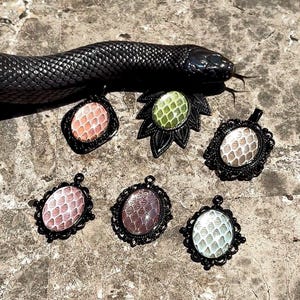 以下が含まれることがあります： 黒い金属製のフレームと、ピンク、緑、白、茶色などの様々な色の質感のあるヘビ革のインサートを備えた6つの楕円形のペンダント。黒いヘビの近くに配置されています。各ペンダントは、滑らかな仕上げの下にユニークな鱗模様が特徴です。
