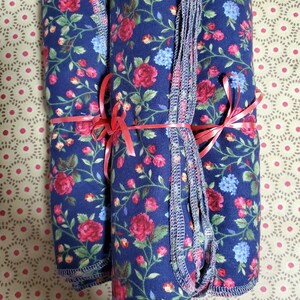 May include: Rolled navy blue fabric with a floral pattern of red roses and blue flowers, tied with red ribbon. The fabric has a decorative stitched edge. The background is a patterned beige surface.