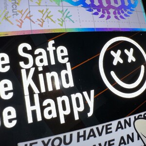 May include: A black car window with a white vinyl sticker that reads "Be Safe Be Kind Be Happy" and a white smiley face with two X's for eyes. The sticker is on the lower right side of the window.