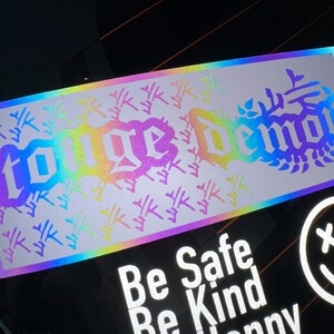 Puede incluir: Una pegatina holográfica con las palabras "Code Demon" en una fuente gótica. La pegatina tiene un fondo degradado arcoíris con un patrón de pequeños árboles estilizados blancos.