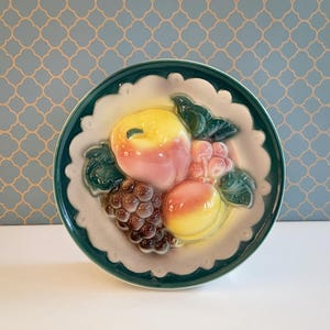 Pode incluir: Um prato de cerâmica com tema de frutas, com borda verde e borda ondulada. O prato apresenta um desenho em relevo de um pêssego, uvas e uma maçã em tons de amarelo, rosa e marrom. O prato tem aproximadamente 20 cm de diâmetro.