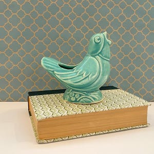 Può includere: Una statuetta di uccello in ceramica color turchese con finitura lucida, caratterizzata da dettagli alari modellati e una piccola cavità aperta sul dorso. L'uccello è appoggiato su una base con motivi a foglie in rilievo sopra un libro.