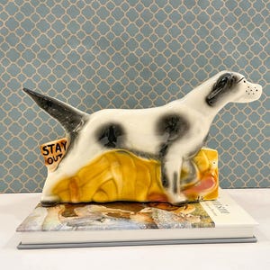 May include: A ceramic dog figurine with a white coat and black spots, standing over a yellow base. The dog has a black tail and ears. A small yellow sign attached to the base features the black text STAY OUT in capital letters.