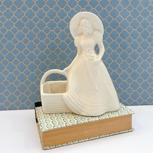 May include: A cream-colored ceramic figurine planter shaped like a person wearing a wide-brimmed hat and a long dress, standing next to a rectangular basket with a handle. The glossy finish reflects light, highlighting the smooth, molded contours.