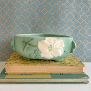 Könnte beinhalten: Hellgrüner Keramik-Blumentopf mit weißem Blumendesign, auf zwei alten Büchern. Der Topf hat eine runde Form mit kleinen Füßen und Griffen. Die Bücher haben vergilbte Einbände und Seiten.