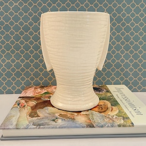 May include: A cream-colored ceramic vase with a textured, horizontal ribbed surface and two prominent, flat, wing-like side handles. The vase features a wide, flared rim and a tapered body that rests on a circular base, showcasing a glossy finish.