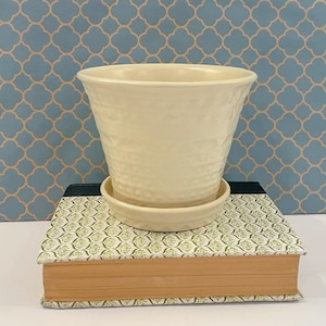 以下が含まれることがあります： 縁が広がったクリーム色の陶器製植木鉢と、お揃いの円形受け皿。鉢の外側には、滑らかで光沢のある仕上げに質感を与える、繊細で繰り返し模様のエンボス加工された幾何学模様が施されています。