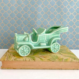 May include: A light green ceramic antique car figurine with spoked wheels and a detailed body. The car sits atop a vintage book with a green and yellow floral design. The background is a light blue wall with a repeating pattern.