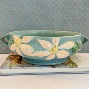Op de afbeelding: Een ronde keramische kom met twee handvatten aan de zijkanten, voorzien van een getextureerd blauwgroen oppervlak versierd met witte bloesems in reliëf met gele harten. De kom heeft een wijde opening en staat op een ronde voet.