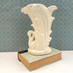 May include: A cream-colored ceramic vase featuring a sculpted swan at the base with elongated, feather-like vertical plumes extending upward to form the rim. The vase is positioned on top of a rectangular book with a patterned cover.