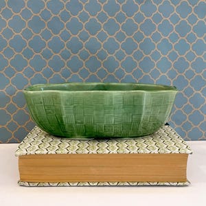 May include: An oval green ceramic bowl with a textured basket-weave pattern on the exterior, featuring a scalloped rim and a glossy finish, resting on top of a thick book with a patterned cover.