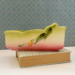 May include: A rectangular ceramic planter with a scalloped rim, painted in a gradient of yellow, pink, and white. The planter features a floral design with green leaves and orange flowers. It rests on a decorative book.