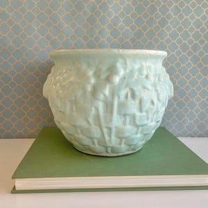 Pode incluir: Um vaso de cerâmica azul claro com um design de tecido de cesta texturizado. O vaso redondo assenta sobre um livro verde contra um fundo azul estampado. O vaso tem duas alças decorativas.